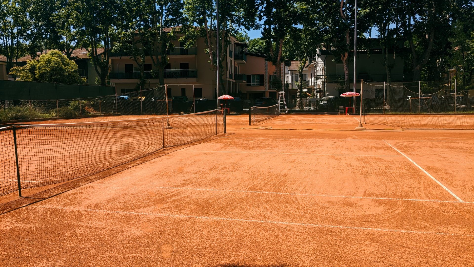Roter Sand-Tennisplatz mit Wohngebäuden und Bäumen im Hintergrund.