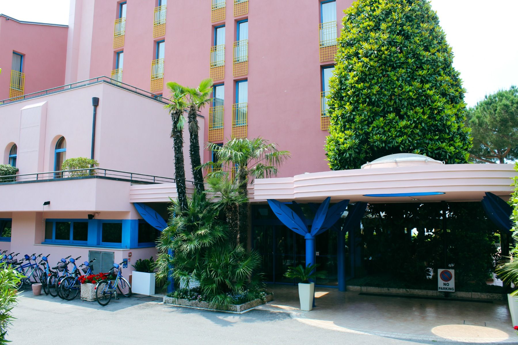 Hoteleingang des Hotel Sportsman am Gardasee mit rosafarbener Fassade, blauen Säulen, Palmen und Leihfahrrädern.