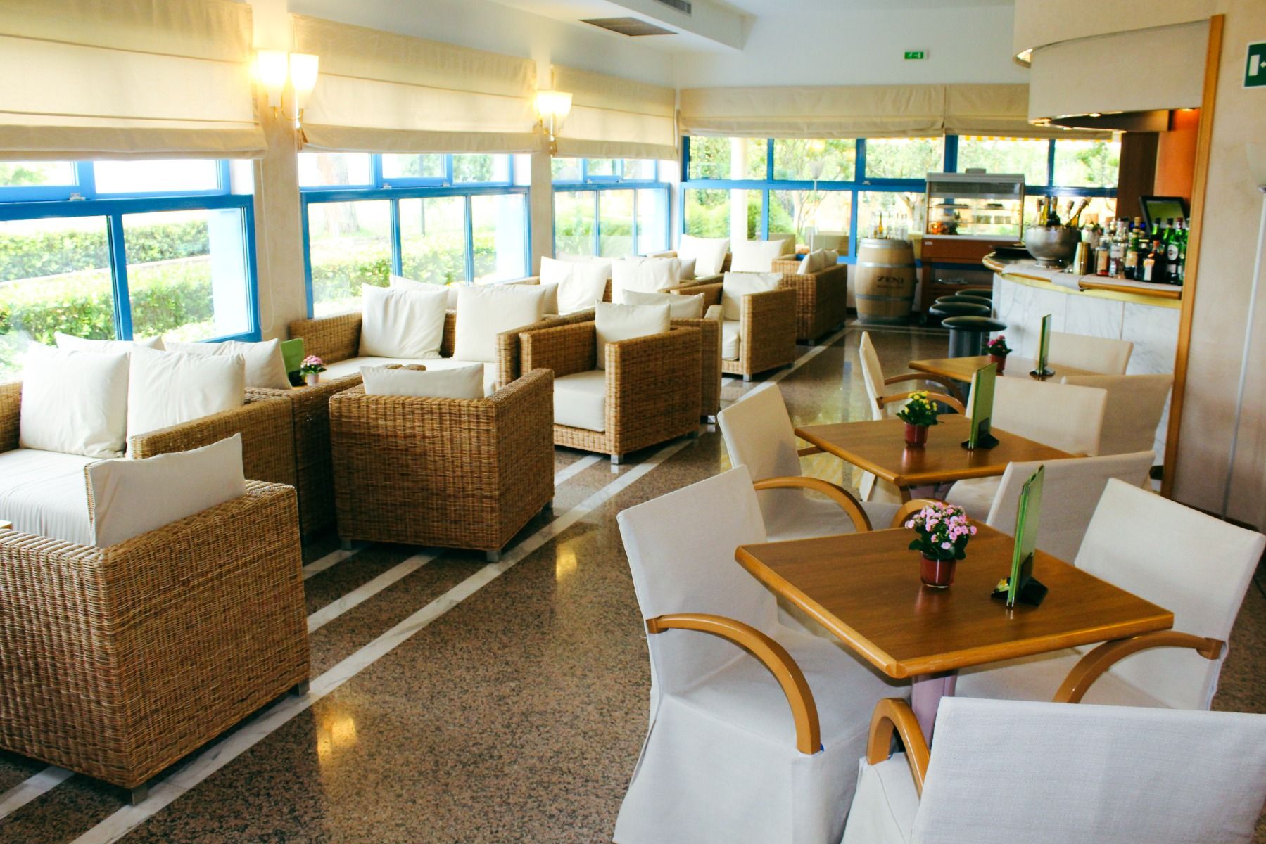 Lounge-Bereich des Hotel Sportsman mit Rattansesseln, Tischen und Bar, großen Fenstern mit Blick ins Grüne.