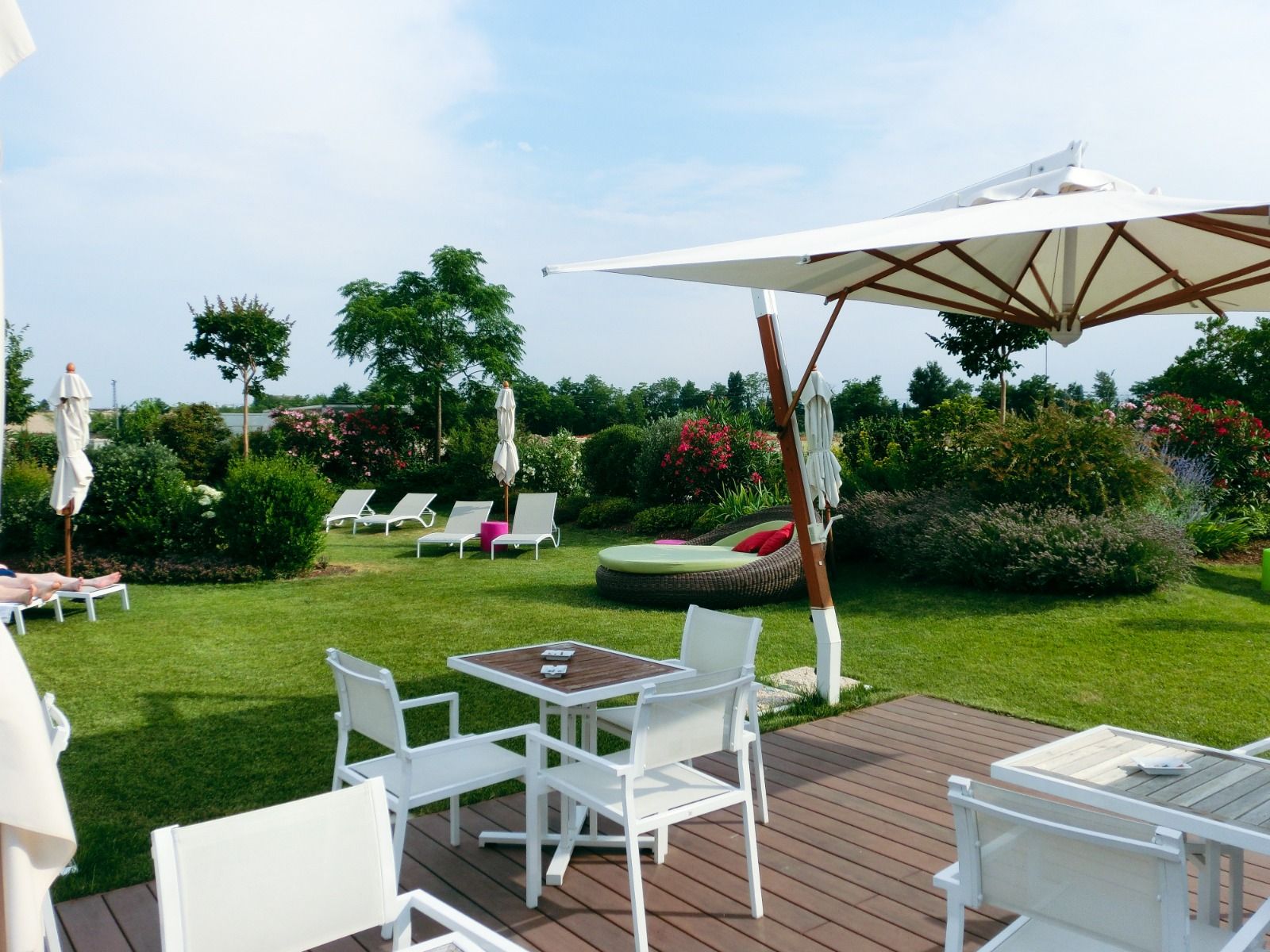 Sonnenterrasse mit weißen Stühlen und Tischen, Sonnenschirmen und grüner Gartenlandschaft im Hintergrund.