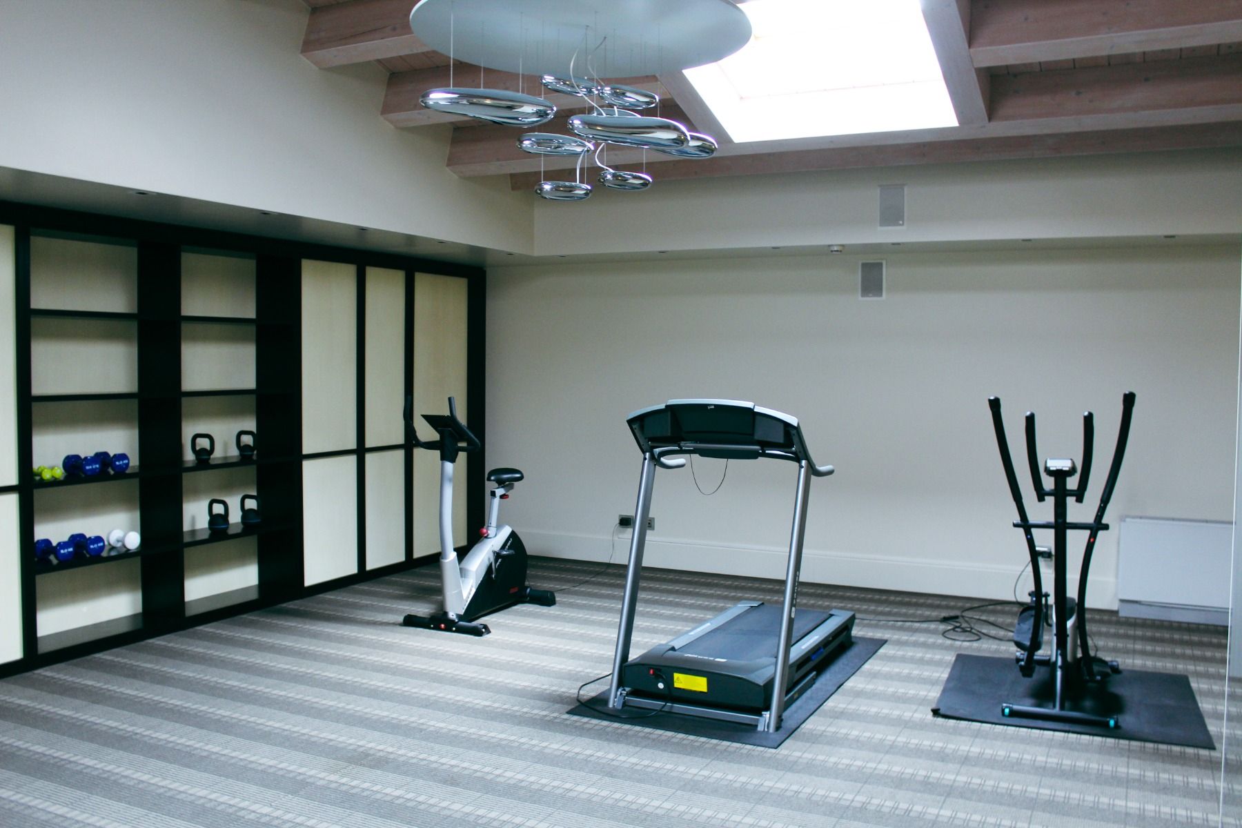 Fitnessraum mit modernen Geräten wie Laufband, Ergometer und Hanteln, helle Atmosphäre mit Dachfenster.