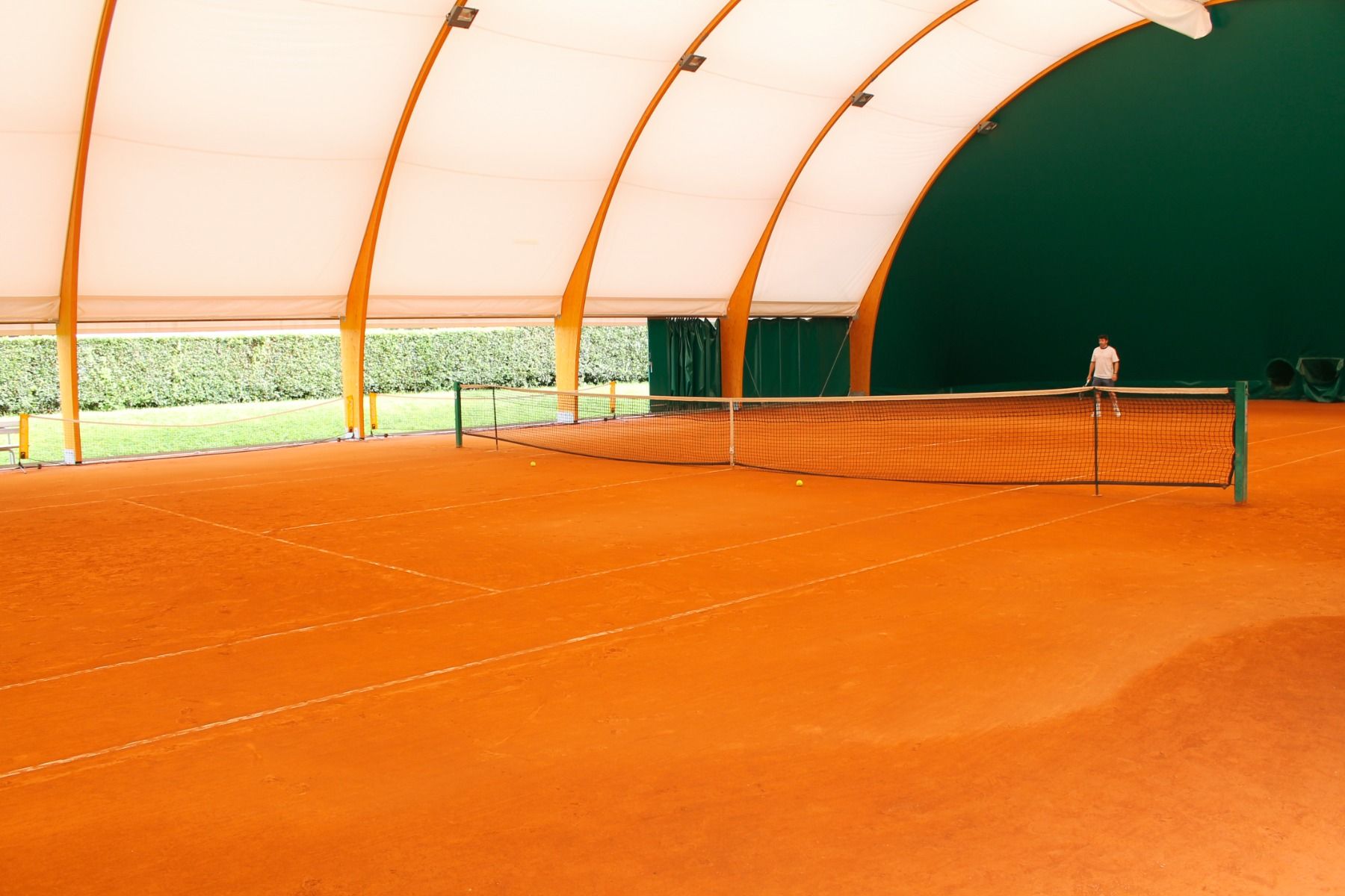 Tennishalle mit rotem Sandplatz und einem Spieler im Hintergrund.