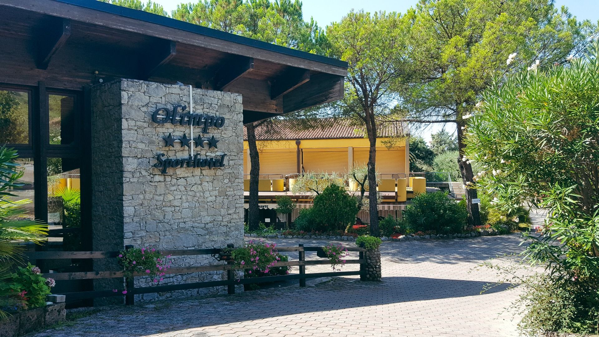 Eingang des Sporthotels Olimpo mit Steinfassade, Hotelname und Sternenmarkierung, umgeben von Pflanzen und Bäumen.