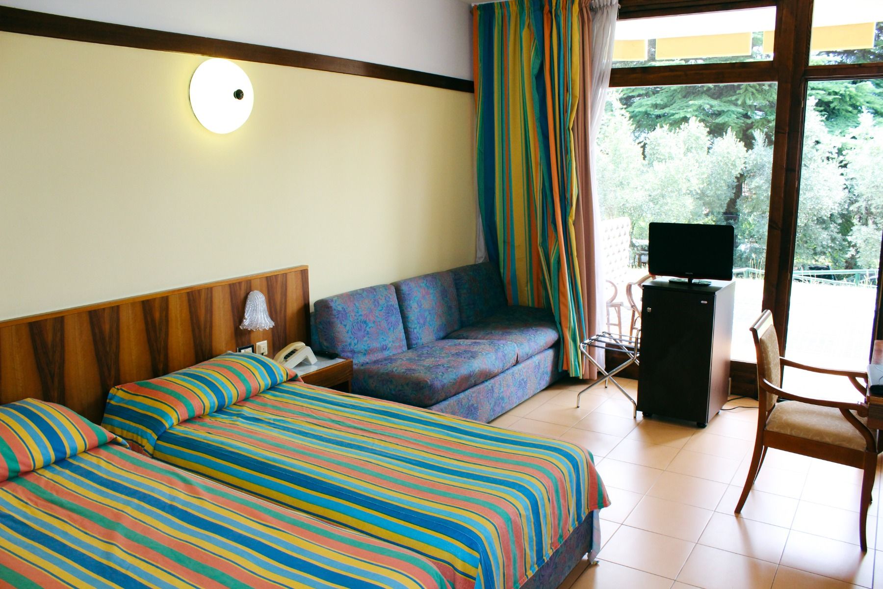Hotelzimmer mit einem Doppelbett, gestreifter Bettwäsche, Sitzbereich und Balkon mit Ausblick ins Grüne.