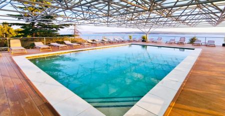 Hinter üppig grünen Bäumen und Sträuchern erhebt sich das hohe, moderne, weiße Gebäude des Hotels Omorika. Eine lange Steintreppe führt hinauf zum Eingang, an dessen Vorderseite ein blaues „Hotel Omorika“-Schild angebracht ist.