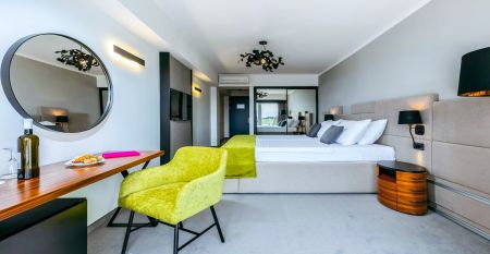 Hinter üppig grünen Bäumen und Sträuchern erhebt sich das hohe, moderne, weiße Gebäude des Hotels Omorika. Eine lange Steintreppe führt hinauf zum Eingang, an dessen Vorderseite ein blaues „Hotel Omorika“-Schild angebracht ist.