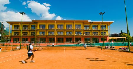 Sand-Tennisplatz mit Spielern, Hotel im Hintergrund und Bergkulisse.