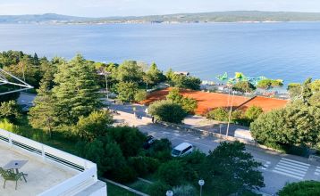 Hinter üppig grünen Bäumen und Sträuchern erhebt sich das hohe, moderne, weiße Gebäude des Hotels Omorika. Eine lange Steintreppe führt hinauf zum Eingang, an dessen Vorderseite ein blaues „Hotel Omorika“-Schild angebracht ist.