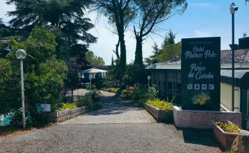 Eingangsbereich des Hotels Marco Polo, von Bäumen und Pflanzen umgeben, mit einem Weg zur Rezeption und einem großen Schild mit Hotelnamen.