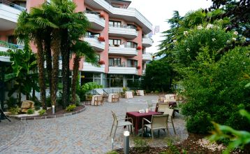 Außenbereich eines Hotels mit rosa Fassade, Balkonen und Palmen, sowie Sitzgelegenheiten im Garten.
