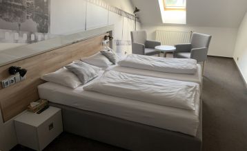 Das Hotel Biograf besticht durch eine hellgelbe und pfirsichfarbene Fassade, auffällige Beschilderungen für das Hotel, das Restaurant und das Café sowie Fenster auf beiden Etagen; links ist ein blaues Straßenschild zu sehen.