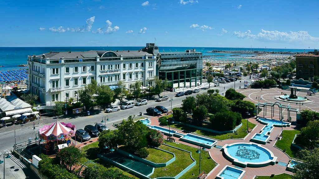 Luftaufnahme der Hotelanlage Kursaal an der Strandpromenade mit Parkanlage, Brunnen und Blick auf das Meer.