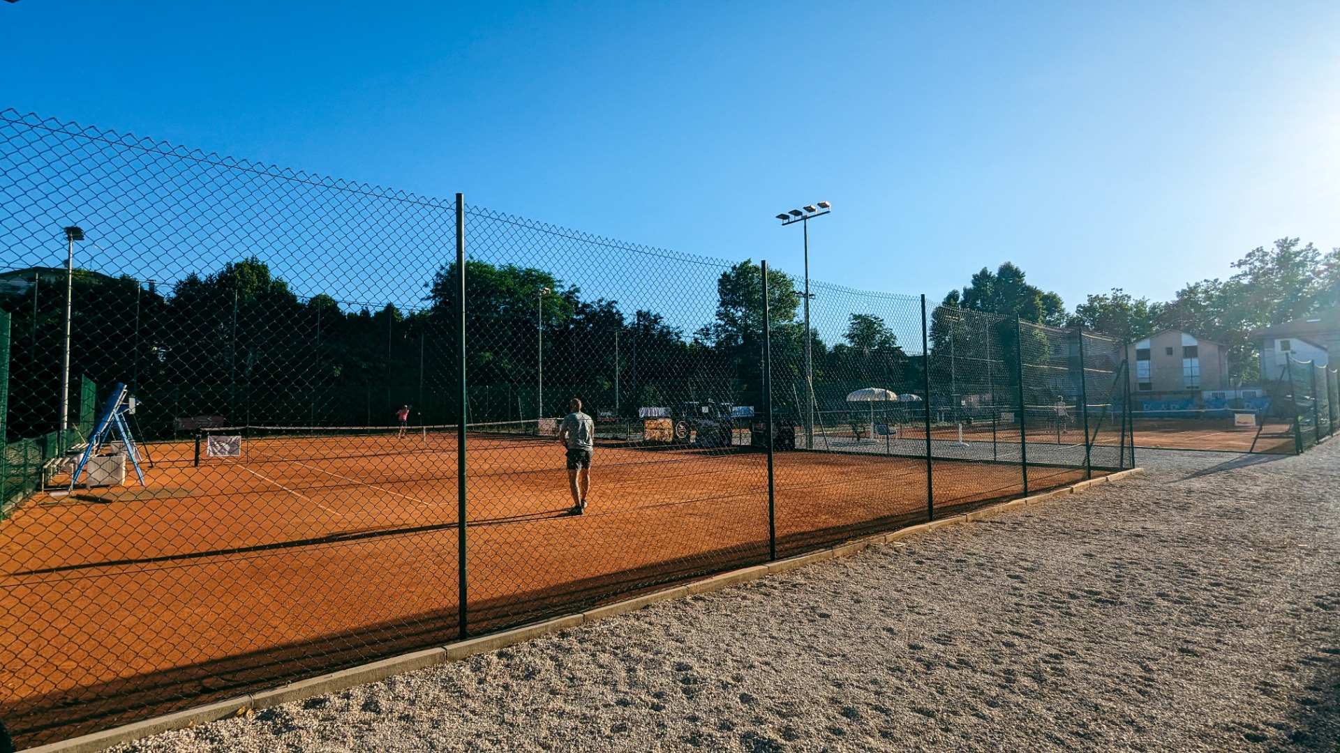 Tennisplatzanlage mit rotem Sandboden, umgeben von Zaun und Flutlicht, zwei Spieler sind auf dem Platz aktiv.